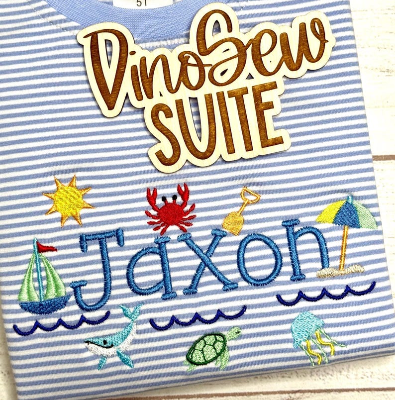 May include: Blue and white striped t-shirt with embroidered beach-themed designs, including a sailboat, sun, crab, and the name "Jaxon" in blue. A wooden cutout reads "DinoSew SUITE".