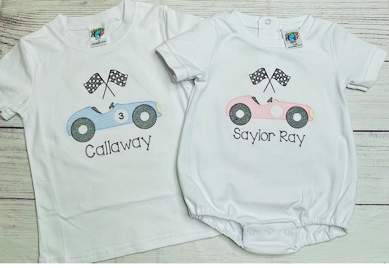 Peut inclure: Deux chemises blanches, l'une un t-shirt et l'autre un body, chacune avec un motif de voiture de course. Le t-shirt a une voiture bleue avec le nom "Callaway" et le num&eacute;ro 3. Le body a une voiture rose avec le nom "Saylor Ray".