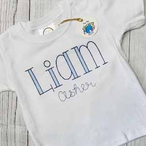 May include: White short-sleeved t-shirt with blue and white striped lettering that says "Liam Asher".