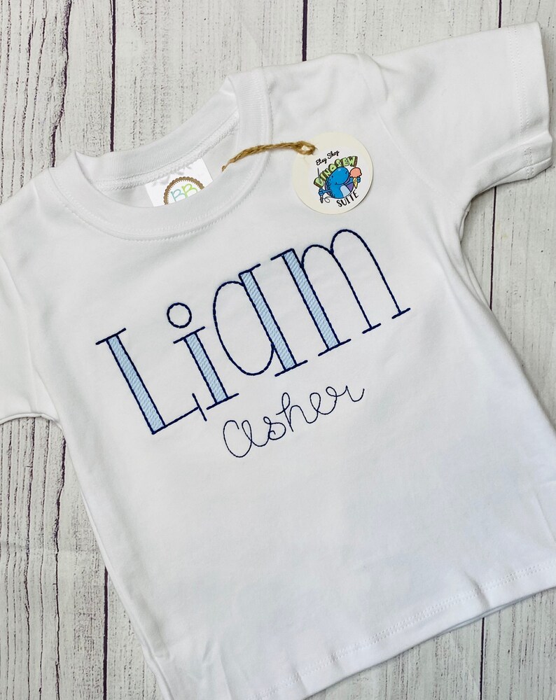 May include: White short-sleeved t-shirt with blue and white striped lettering that says "Liam Asher".