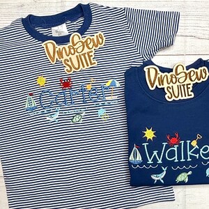 May include: Two children's shirts. One is navy blue with the name "Walker" embroidered in blue, and the other is striped with the name "Carter". Both shirts have beach-themed embroidery, including a sailboat, sun, and crab.