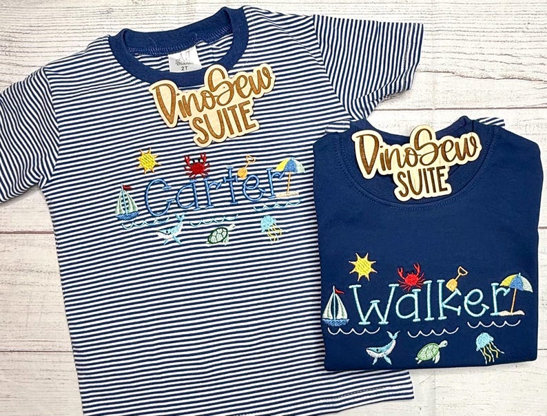 May include: Two children's shirts. One is navy blue with the name "Walker" embroidered in blue, and the other is striped with the name "Carter". Both shirts have beach-themed embroidery, including a sailboat, sun, and crab.