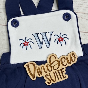 May include: Navy blue baby overalls with white bib. The bib features a blue embroidered letter "W" with red and blue firework designs. The overalls have navy blue straps and buttons. A wooden sign says "DinoSew SUITE".