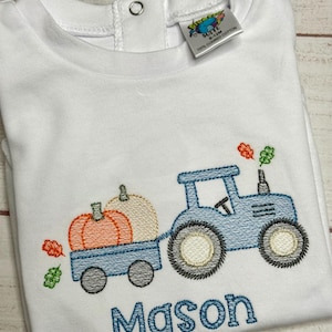 May include: A white long-sleeve shirt featuring an embroidered blue tractor pulling a trailer with orange and cream pumpkins. The design includes small orange and green leaves and the name Mason in blue thread. A tag indicates 100% combed cotton.