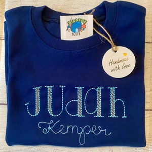 May include: A navy blue t-shirt with the name "JUDAH Kemper" embroidered in blue and silver thread. The shirt has a tag that says "Handmade with love".