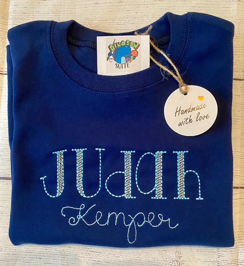 May include: A navy blue t-shirt with the name "JUDAH Kemper" embroidered in blue and silver thread. The shirt has a tag that says "Handmade with love".