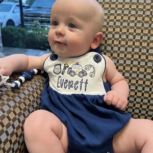 May include: A baby boy wearing a blue and white onesie with a police car and other police-themed embroidery. The onesie says "Everett" on the front.