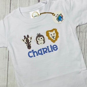 May include: White short-sleeved t-shirt with embroidered animal faces and the name "charlie" in blue thread.
