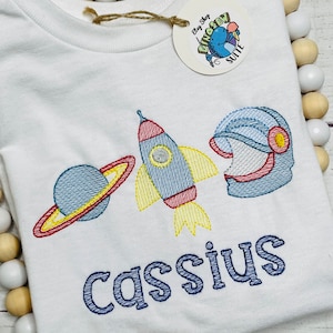 May include: White t-shirt with embroidered space-themed design, including a rocket, planet, and astronaut helmet. The name "Cassius" is embroidered in blue below the design.