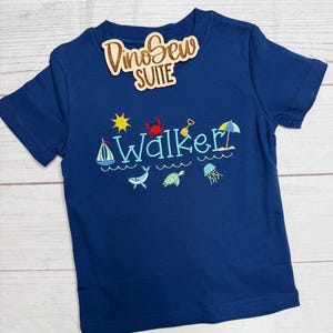 May include: Navy blue short-sleeved t-shirt with embroidered beach-themed designs, including a sailboat, crab, and sun. The name "Walker" is embroidered in light blue. A wooden tag with the text "DinoSew SUITE" is attached.