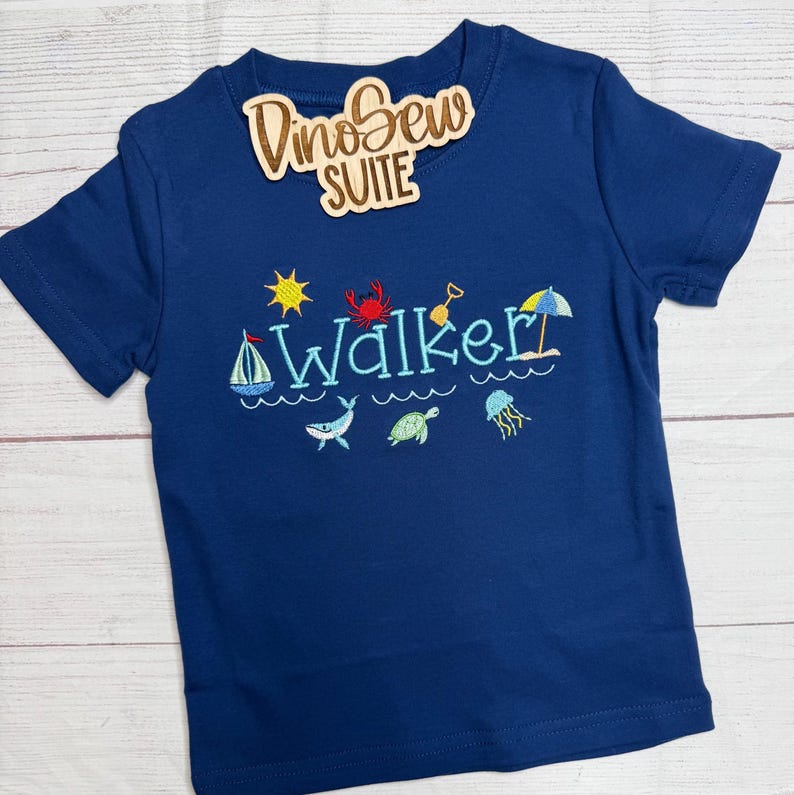 May include: Navy blue short-sleeved t-shirt with embroidered beach-themed designs, including a sailboat, crab, and sun. The name "Walker" is embroidered in light blue. A wooden tag with the text "DinoSew SUITE" is attached.