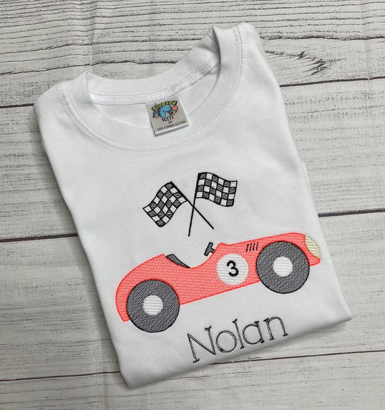 Peut inclure: T-shirt blanc avec une broderie d'une voiture de course corail, le num&eacute;ro 3 et le nom "Nolan". Deux drapeaux &agrave; damiers sont au-dessus de la voiture. Le t-shirt porte une &eacute;tiquette "SUITE".