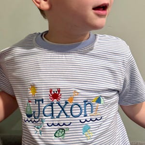 May include: A light blue and white striped t-shirt with the name "Jaxon" embroidered in blue. The shirt features beach-themed embroidery, including a sun, crab, sailboat, and waves. The shirt has a light blue collar.