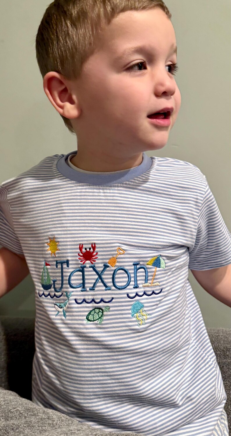 May include: A light blue and white striped t-shirt with the name "Jaxon" embroidered in blue. The shirt features beach-themed embroidery, including a sun, crab, sailboat, and waves. The shirt has a light blue collar.