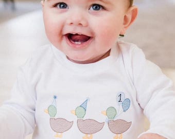 May include: A baby wearing a white long-sleeved onesie with three embroidered ducks wearing party hats and a balloon. The onesie has the name "Chip" embroidered on the bottom. The baby is wearing a green felt crown with a wooden number "1" on it.