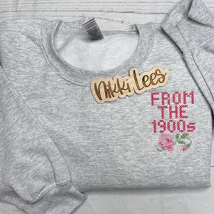 May include: A heather gray sweatshirt with the text "FROM THE 1900s" in pink cross-stitch font, along with a floral design. A wooden sign that says "Nikki Lees" is on top of the sweatshirt.