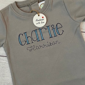 May include: A gray long-sleeve baby onesie with blue and white striped embroidery that reads "Charlie Harrison". A small wooden tag with a red heart and the text "Handmade with love" is attached to the onesie.