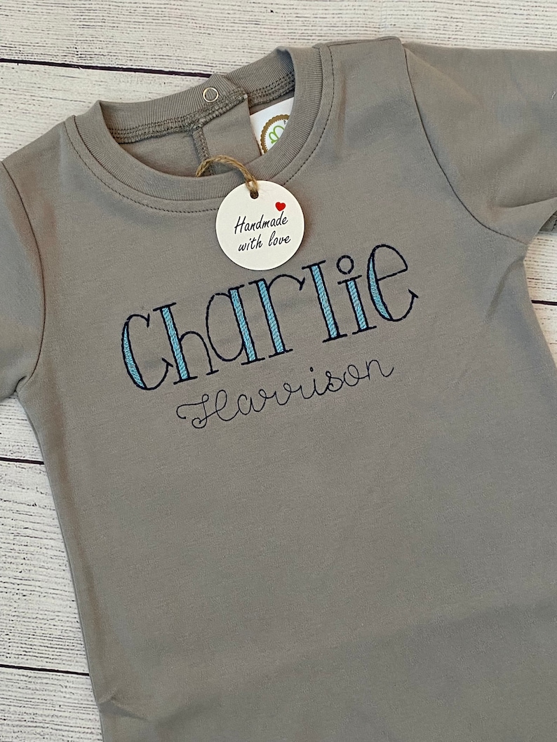 May include: A gray long-sleeve baby onesie with blue and white striped embroidery that reads "Charlie Harrison". A small wooden tag with a red heart and the text "Handmade with love" is attached to the onesie.