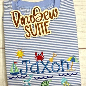 May include: A light blue and white striped t-shirt with the name "Jaxon" embroidered in blue, along with beach-themed designs. A wooden sign that says "DinoSew SUITE" is on top of the shirt.