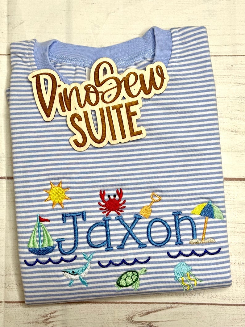 May include: A light blue and white striped t-shirt with the name "Jaxon" embroidered in blue, along with beach-themed designs. A wooden sign that says "DinoSew SUITE" is on top of the shirt.