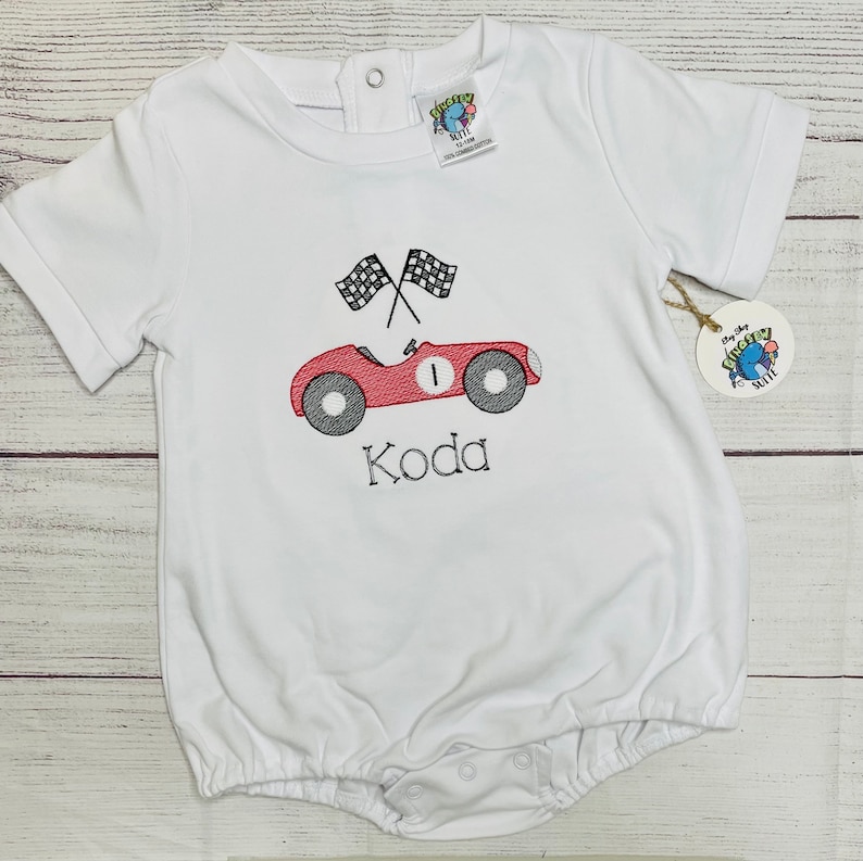 Peut inclure: Barboteuse blanche pour b&eacute;b&eacute; avec un motif de voiture de course rouge, des drapeaux &agrave; damier noirs et blancs, et le nom "Koda" brod&eacute; en noir. La barboteuse a des manches courtes et des fermetures &agrave; boutons-pression.