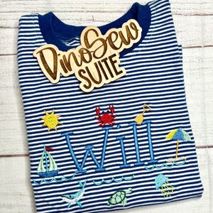 May include: A navy blue and white striped long-sleeve shirt with embroidered beach-themed designs, including a sailboat, sun, crab, and the name "WILL". A wooden sign that says "Dino Sew SUITE" is on top.