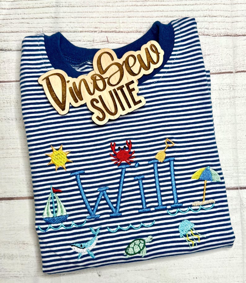 May include: A navy blue and white striped long-sleeve shirt with embroidered beach-themed designs, including a sailboat, sun, crab, and the name "WILL". A wooden sign that says "Dino Sew SUITE" is on top.