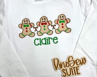 May include: White long-sleeve shirt featuring three gingerbread man designs with red and green accents. The name "Claire" is embroidered in green below the gingerbread men. A wooden sign with the text "DinoSew SUITE" is also visible.