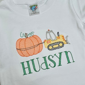 May include: A white t-shirt with an embroidered design of a pumpkin, a yellow construction vehicle, and the name "HUDSYN" in green.