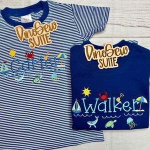 May include: Two children's t-shirts. One is navy blue, the other is white and navy blue striped. Both shirts have embroidered beach-themed designs, including a sailboat, sun, crab, and the names "Carter" and "Walker" in blue. Each shirt has a wooden tag that says "DinoSew SUITE".