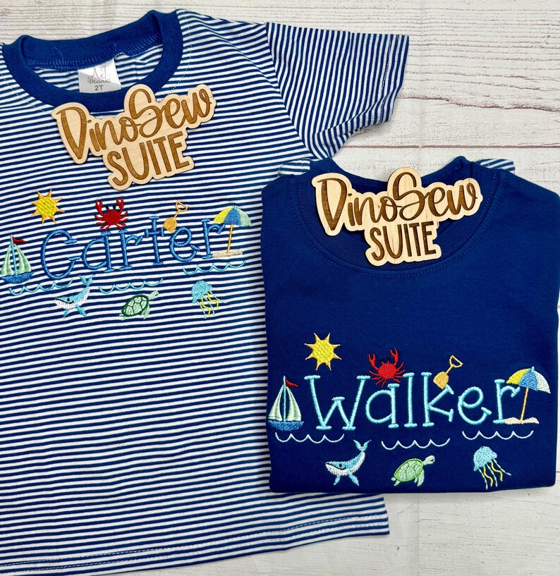 May include: Two children's t-shirts. One is navy blue, the other is white and navy blue striped. Both shirts have embroidered beach-themed designs, including a sailboat, sun, crab, and the names "Carter" and "Walker" in blue. Each shirt has a wooden tag that says "DinoSew SUITE".