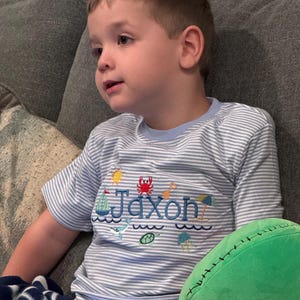 May include: A young child wearing a light blue and white striped t-shirt with the name "Jaxon" embroidered on the front. The shirt features nautical-themed embroidery, including a sailboat, crab, and other sea creatures.