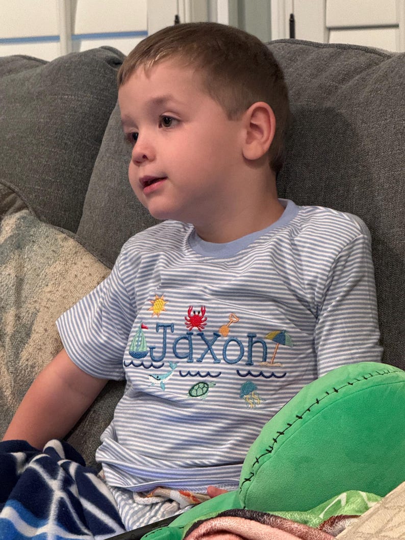 May include: A young child wearing a light blue and white striped t-shirt with the name "Jaxon" embroidered on the front. The shirt features nautical-themed embroidery, including a sailboat, crab, and other sea creatures.