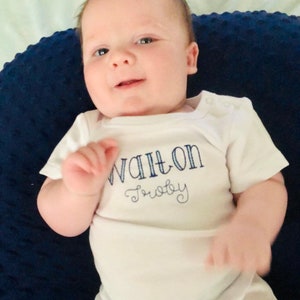 May include: A white baby onesie with blue embroidery that reads "Walton Baby". The onesie is worn by a baby lying on a blue and white patterned pillow.