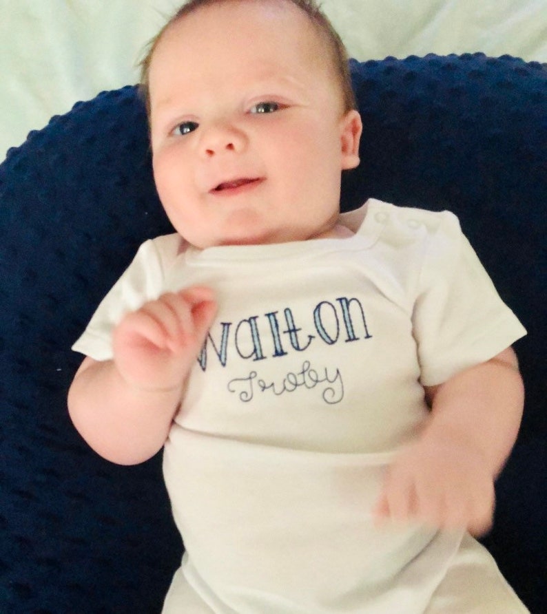 May include: A white baby onesie with blue embroidery that reads "Walton Baby". The onesie is worn by a baby lying on a blue and white patterned pillow.
