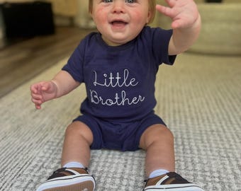 May include: A smiling baby wearing a navy blue t-shirt and shorts set with the words "Little Brother" in white script. The baby is wearing brown, white, and navy blue sneakers with the brand name "Sperry" on the sole.