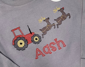 Puede incluir: Camiseta gris de manga larga con un tractor rojo tirando de dos renos con cuernos amarillos. El nombre "Aash" está bordado en rojo. Una etiqueta con un logotipo está sujeta a la camisa.