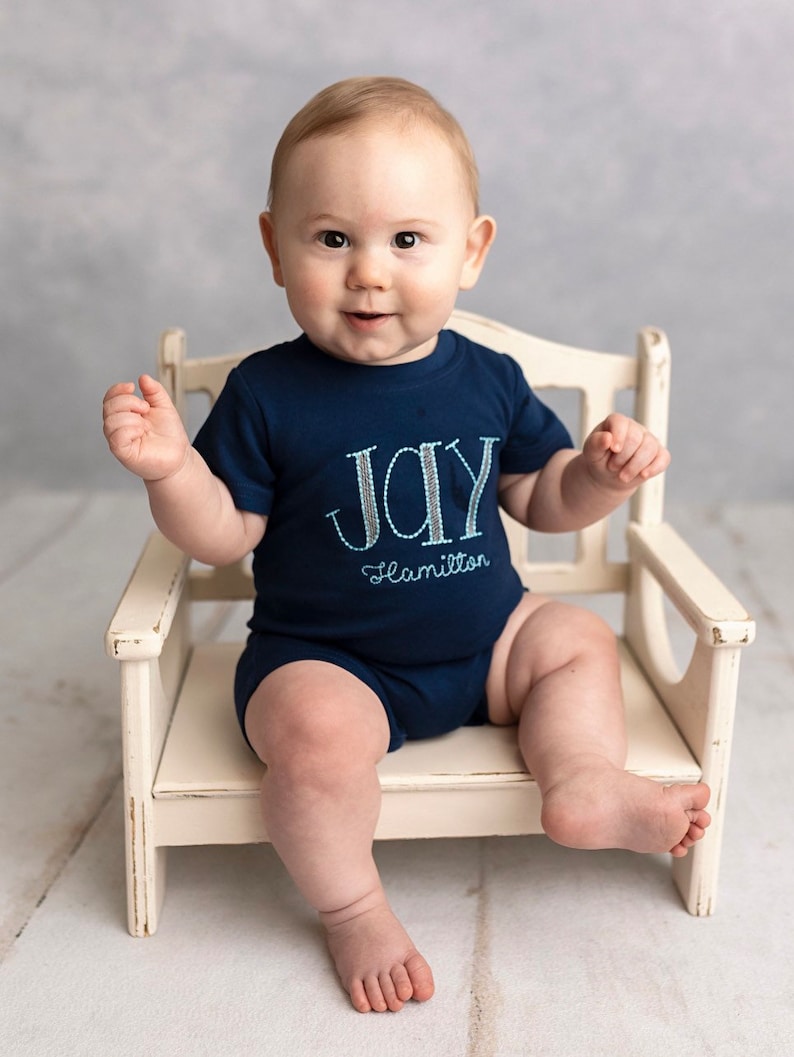 May include: A baby boy wearing a blue onesie with the word "JOY" and the name "Hamilton" embroidered on it. The baby is sitting on a white wooden chair.