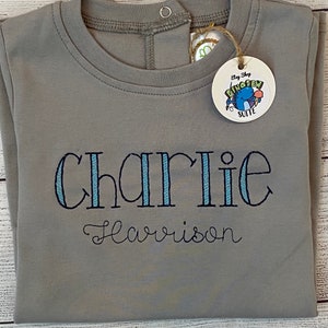 May include: Gray long-sleeve shirt with a snap closure at the neck. The shirt is embroidered with the name "Charlie Harrison" in blue and white thread. A tag with the text "Etsy Shop Dino Sew Suite" is attached to the shirt.