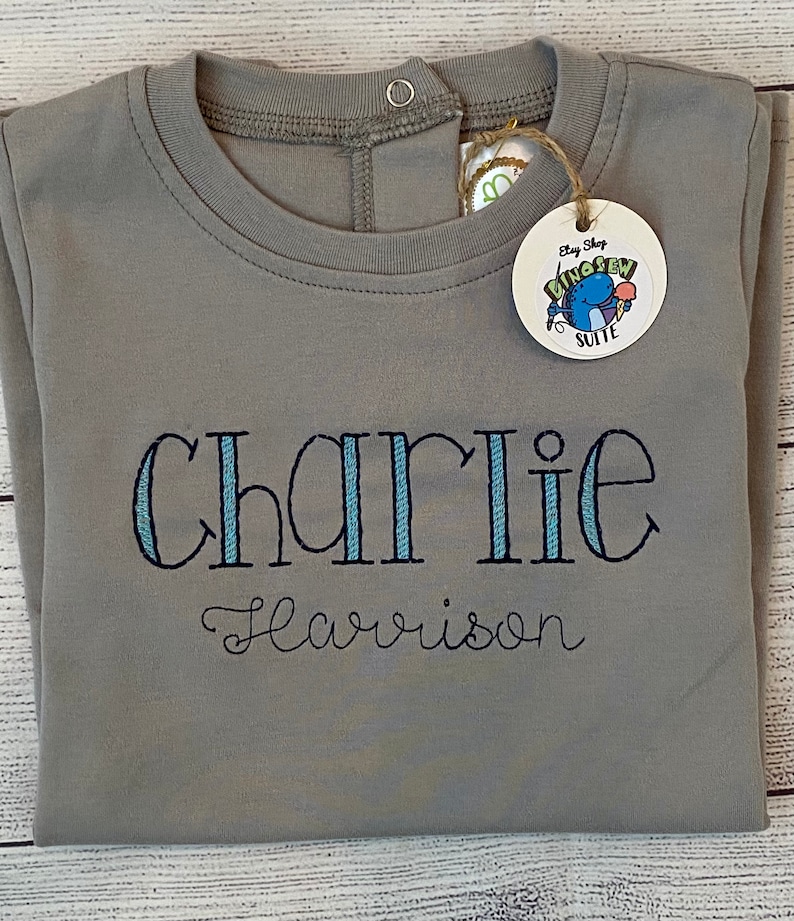 May include: Gray long-sleeve shirt with a snap closure at the neck. The shirt is embroidered with the name "Charlie Harrison" in blue and white thread. A tag with the text "Etsy Shop Dino Sew Suite" is attached to the shirt.