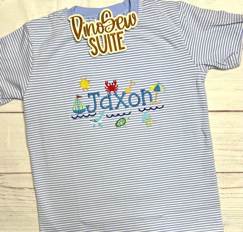 May include: A light blue and white striped t-shirt with the name "Jaxon" embroidered in blue, along with beach-themed designs like a sailboat, sun, crab, and waves. The shirt also has the text "DinoSew SUITE" on a tag.