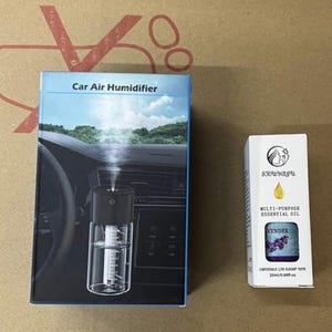May include: A box labeled "Car Air Humidifier" next to a small white box of "Multi-Purpose Essential Oil". The car humidifier box shows a black and clear humidifier inside a car. The essential oil box has a lavender design.