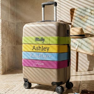 Op de afbeelding: Een beige rolkoffer met vier gekleurde banden met de namen Molly, Ashley, Kaitlyn en Jillian. Een hoed en geld liggen op de grond in de buurt van een stoel.