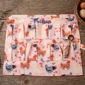May include: Pink egg gathering apron with a chicken and floral pattern. The apron has multiple pockets for collecting eggs and the name "Melissa" embroidered in blue. A wooden spoon rests on the apron.