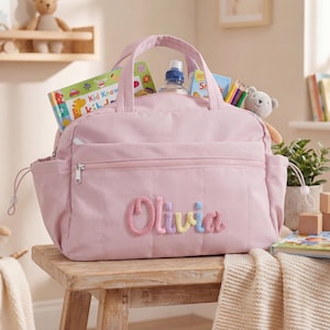 Op de afbeelding: Een lichtroze luiertas met een handvat en zijvakken, gepersonaliseerd met de naam "Olivia" in kleurrijke geborduurde letters. De tas bevat boeken, een waterfles en een klein knuffeldier. Hij staat op een houten krukje.