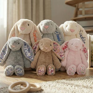 May include: A collection of plush bunny rabbits in various colors, including gray, beige, and pink. The bunnies have long ears with floral patterned fabric and embroidered names. The bunnies are sitting on a wooden floor.