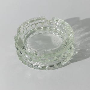May include: A clear glass ashtray with a circular design. The ashtray has a textured, geometric pattern around the rim and a smooth interior. The ashtray is designed to hold cigarettes or cigars and is suitable for home or office use.