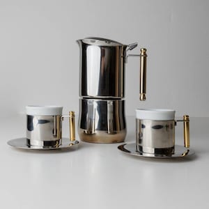 Könnte beinhalten: Eine Kaffeemaschine aus Edelstahl mit goldenem Griff und zwei passenden Tassen und Untertassen. Die Tassen haben weiße Innenräume und goldene Griffe. Das Set ist zum Brühen und Servieren von Kaffee bestimmt.