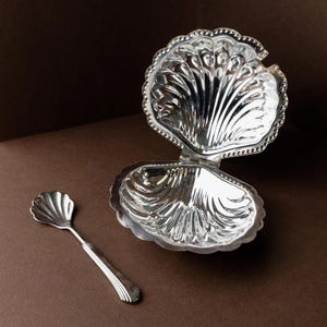 Puede incluir: Un plato plateado en forma de concha con tapa adjunta y una cuchara pequeña. El plato está abierto, revelando el interior. La cuchara y el plato tienen un diseño detallado y acanalado. El fondo es de color marrón liso.