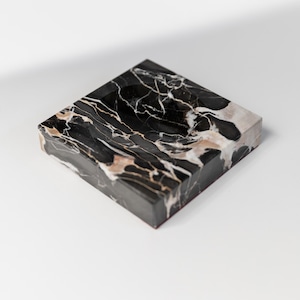 Könnte beinhalten: Quadratische schwarze Marmorbox mit weißen und beigen Adern. Die Box hat eine polierte Oberfläche und einen roten Sockel. Das Marmormuster bildet einen auffälligen Kontrast zum schwarzen Hintergrund. Diese dekorative Box ist ideal für die Aufbewahrung kleiner Gegenstände.